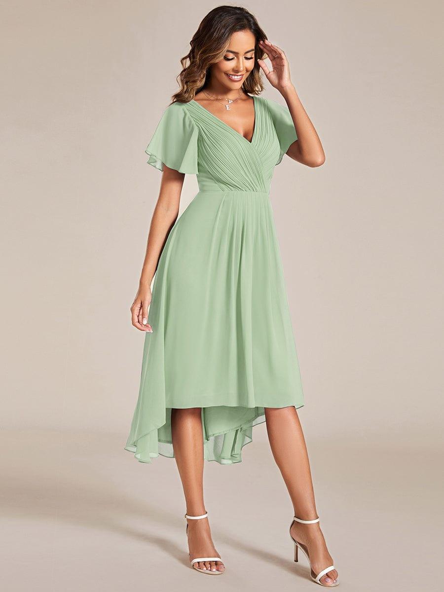 Salbeigrünes Chiffonkleid in seitlicher Dreiviertelansicht, Model berührt lächelnd ihr Haar, Saum vorn kürzer und hinten länger. 80