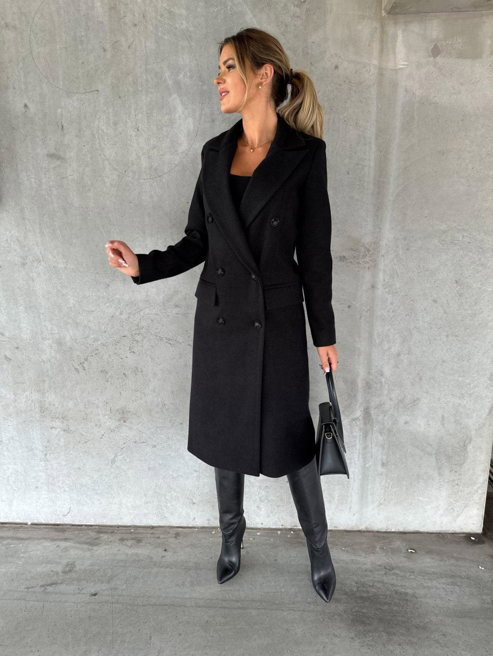 Frau im eleganten schwarzen Wollmantel, trägt Stiefel und Handtasche, vor Betonwand.