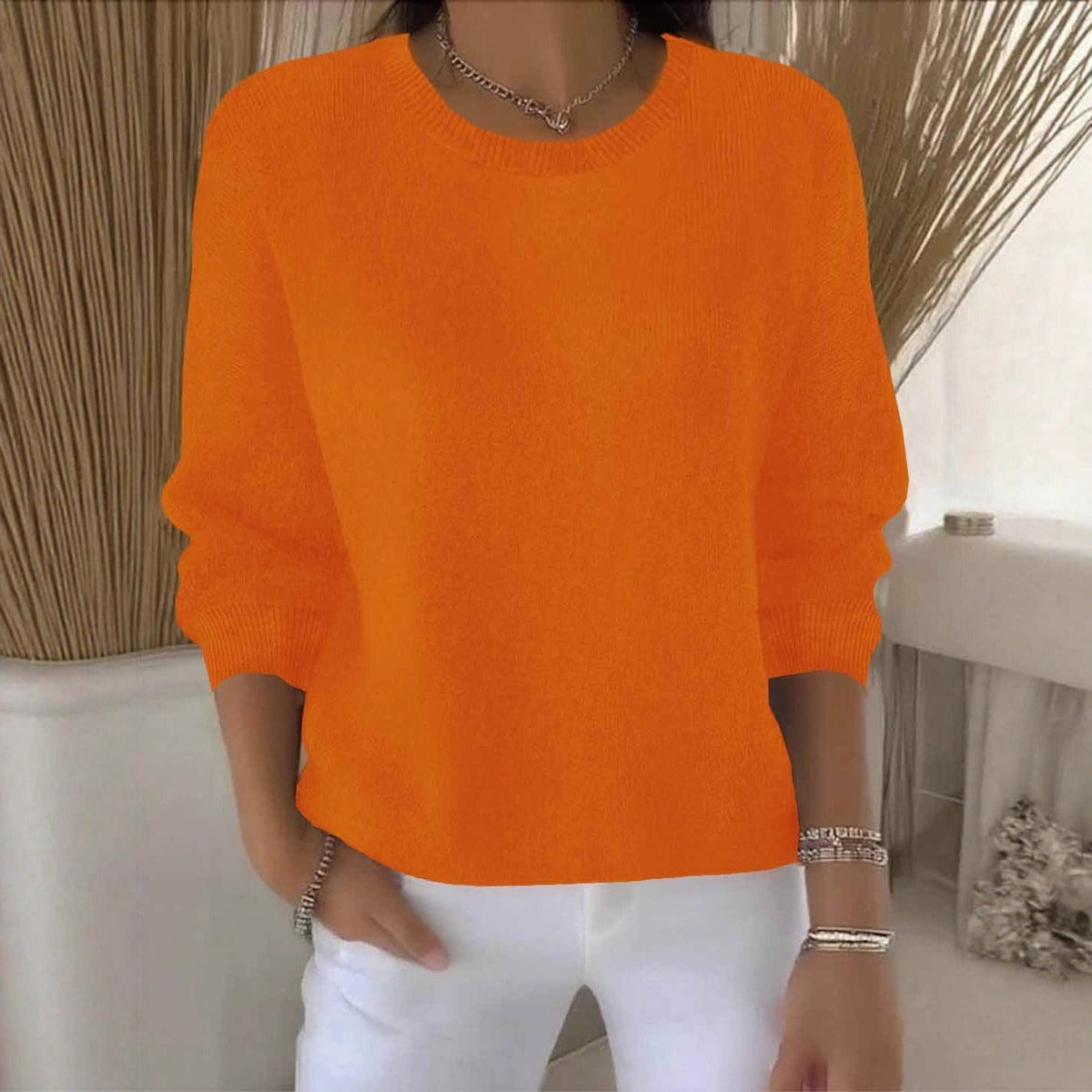Model im orangefarbenen Strickpullover mit Rundhals, weiße Hose, Dekoration aus Trockengras im Hintergrund 11