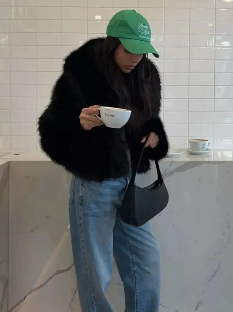 Frau im schwarzen Kunstpelzmantel mit grüner Kappe und Jeans hält in einem Café eine Tasse 10