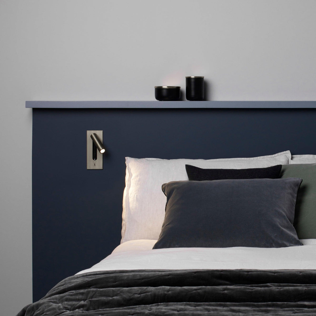 Bronzefarbene Leseleuchte an blauem Kopfteil über einem Bett mit grauen Kissen und Regalbrett 30