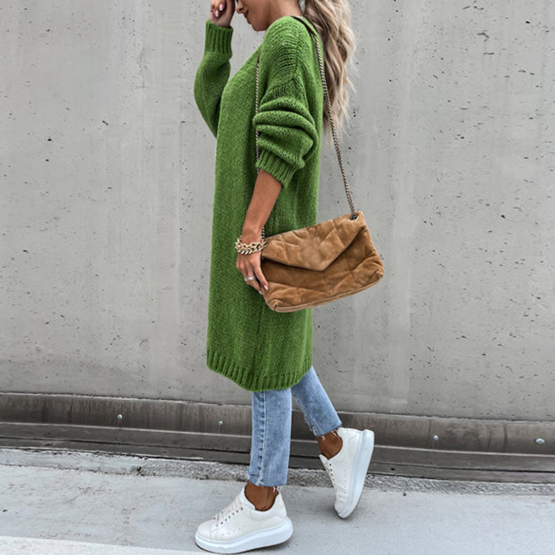 Frau in grünem Strickkleid, blauer Jeans, weißen Sneakers, braune Umhängetasche, Betonhintergrund.