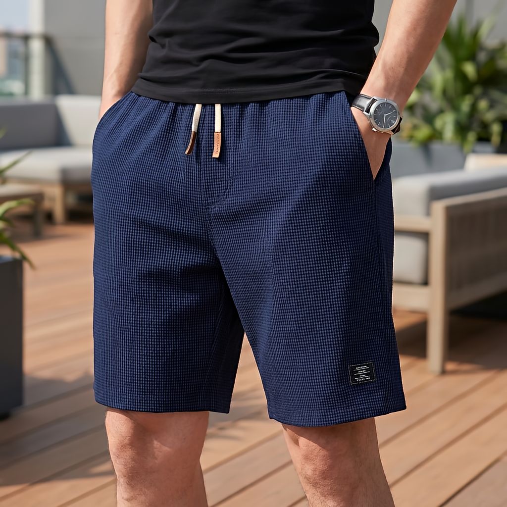 Model trägt die Shorts in Marineblau mit Waffelstruktur und hellem Kordelzug, Hand in der Tasche, auf einer sonnigen Dachterrasse. 7