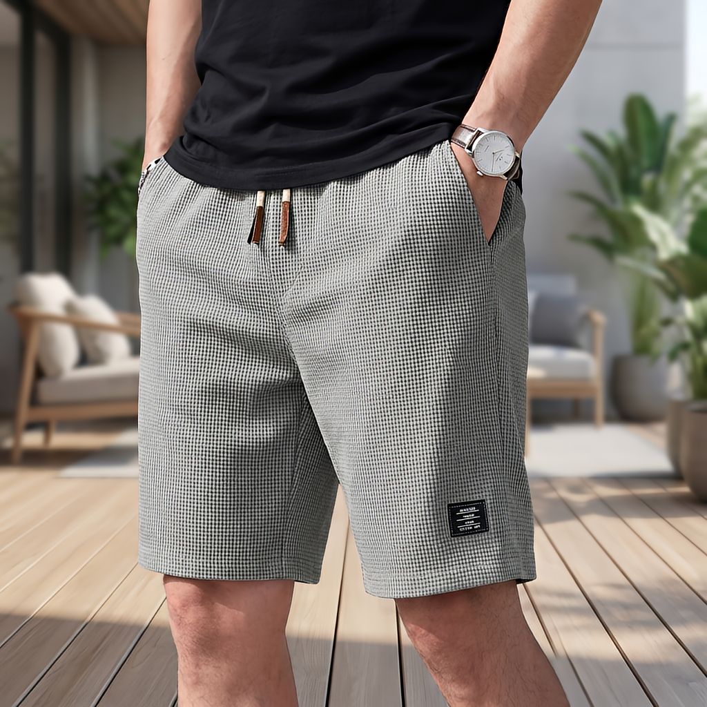 Model in grauen Shorts mit feiner Waffelstruktur und Kordelzug, eine Hand in der Tasche, dazu Armbanduhr und schwarzes T-Shirt. 6