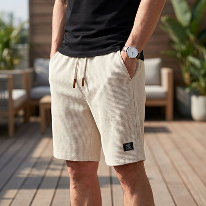 Model in hellbeigen Shorts mit Waffelstruktur und Kordelzug, Hand in der Seitentasche, auf einer Holzterrasse mit Sitzmöbeln. 8