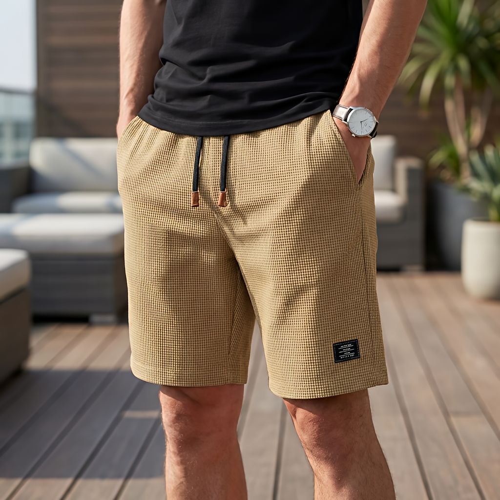 Model in sandfarbenen Shorts mit Waffelstruktur und dunklem Kordelzug, Hand in der Tasche, vor Loungemöbeln auf einer Holzterrasse. 4