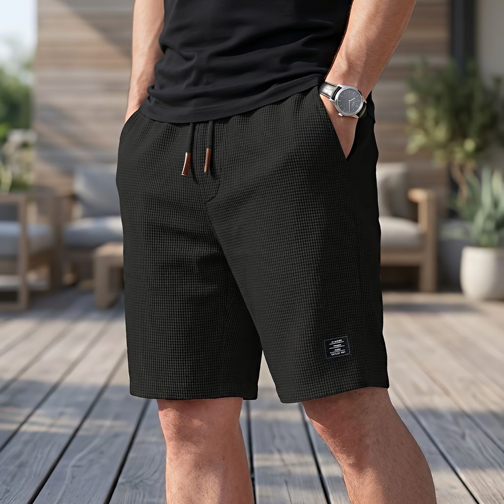 Model trägt die Shorts in Schwarz mit Waffelmuster und Kordelzug, eine Hand in der Tasche, dazu Armbanduhr und schwarzes T-Shirt. 3