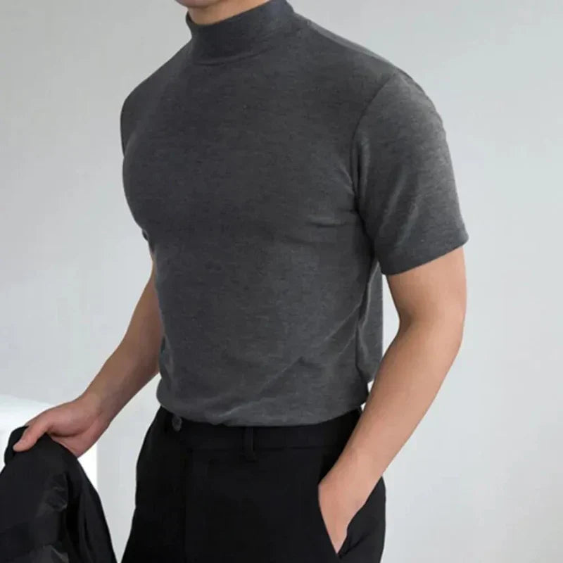 Mann im dunkelgrauen Kurzarmshirt mit Stehkragen, eine Hand in der Hosentasche der schwarzen Hose, in der anderen Hand ein dunkles Kleidungsstück. 3