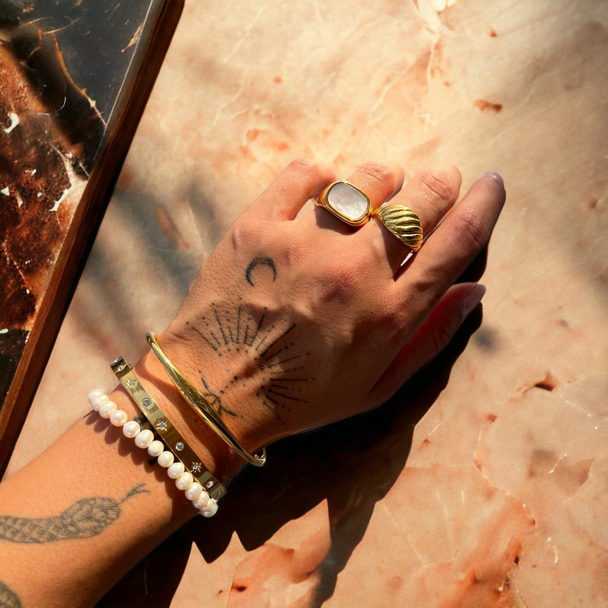 Lysa Goldarmband am Handgelenk mit weiteren Armbändern und Ringen auf rosa Marmor, another view