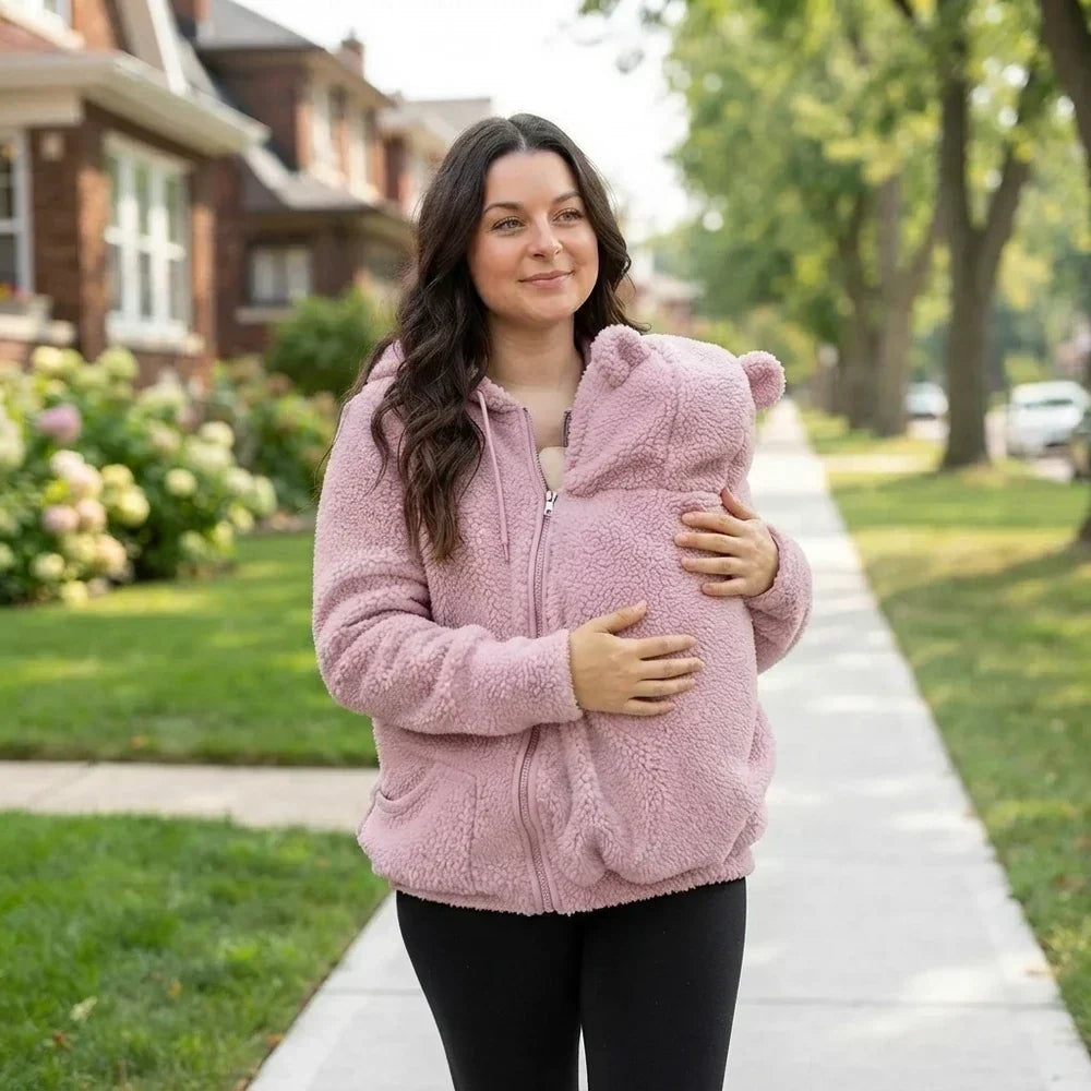 Frau in rosa Teddyfleece-Pullover mit Bärenohren-Kapuze spaziert und trägt ihr Baby geborgen vor der Brust.