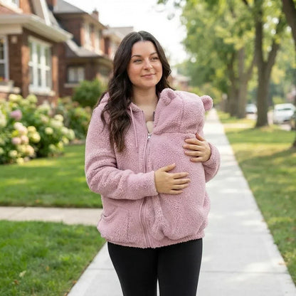 Frau in rosa Teddyfleece-Pullover mit Bärenohren-Kapuze spaziert und trägt ihr Baby geborgen vor der Brust.
