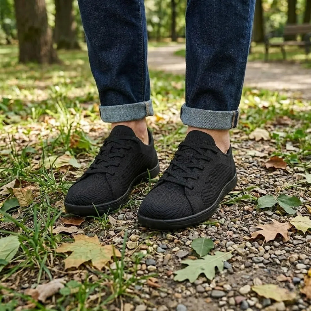 Schwarze Sneaker an den Füßen einer Person in hochgekrempelter Jeans auf einem Waldweg mit Kies und Herbstlaub. 11
