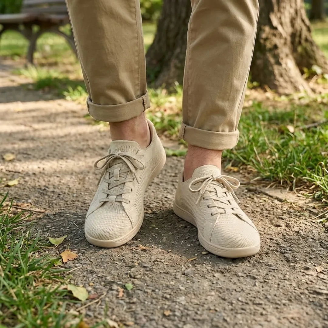 Beige Sneaker an den Füßen einer Person mit hochgekrempelter Hose, die auf einem Parkweg zwischen Gras und Laub steht. 2