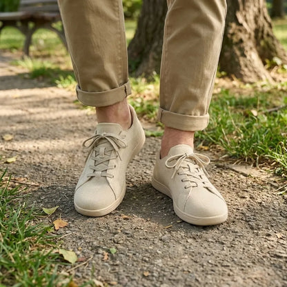 Beige Sneaker an den Füßen einer Person mit hochgekrempelter Hose, die auf einem Parkweg zwischen Gras und Laub steht. 2