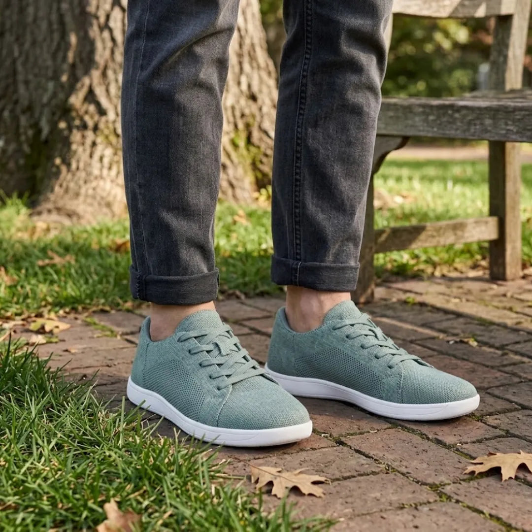 Person in dunkler Jeans trägt die graugrünen Sneaker mit weißer Sohle auf Backsteinpflaster neben Wiese und Parkbank 29