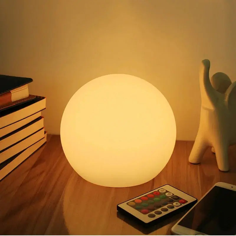 Wiederaufladbare LED-Kugellampe in Weiß auf einem Holztisch neben Büchern, einer Keramikfigur, der Fernbedienung und einem Smartphone.