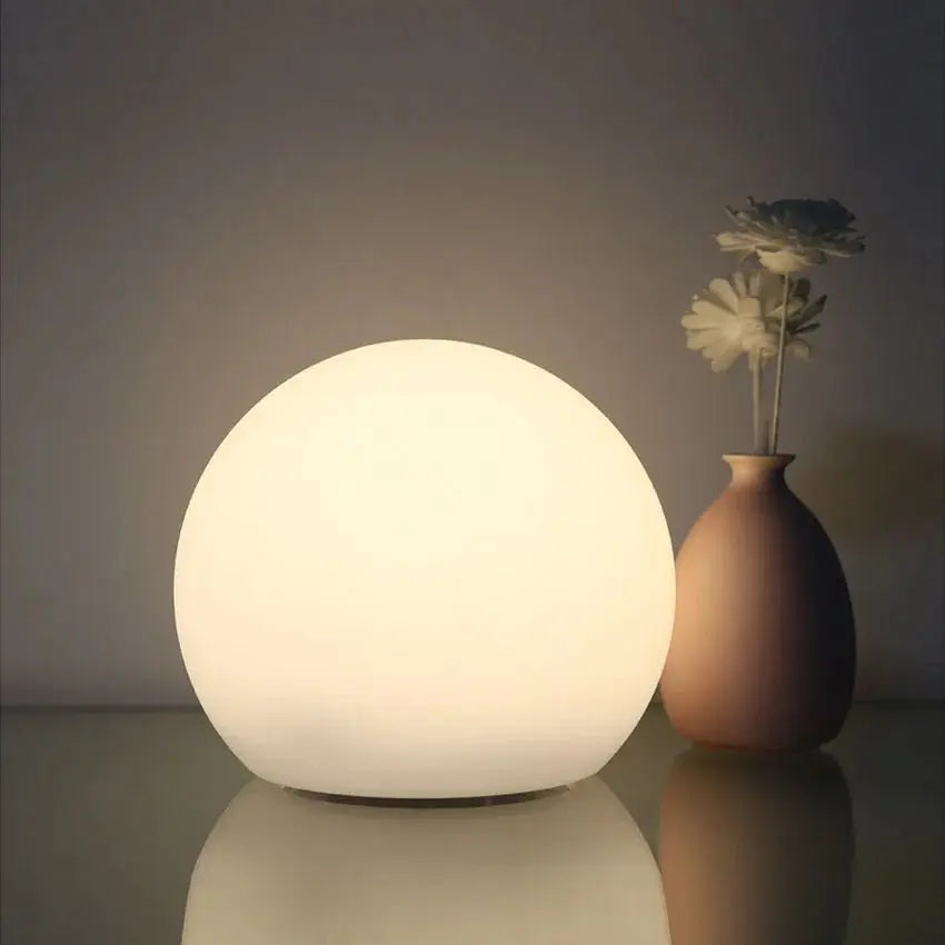 Wiederaufladbare LED-Kugellampe in Weiß auf einer spiegelnden Fläche neben einer braunen Vase mit Blumen.