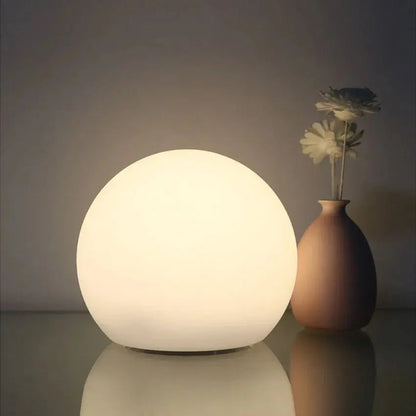 Wiederaufladbare LED-Kugellampe in Weiß auf einer spiegelnden Fläche neben einer braunen Vase mit Blumen.