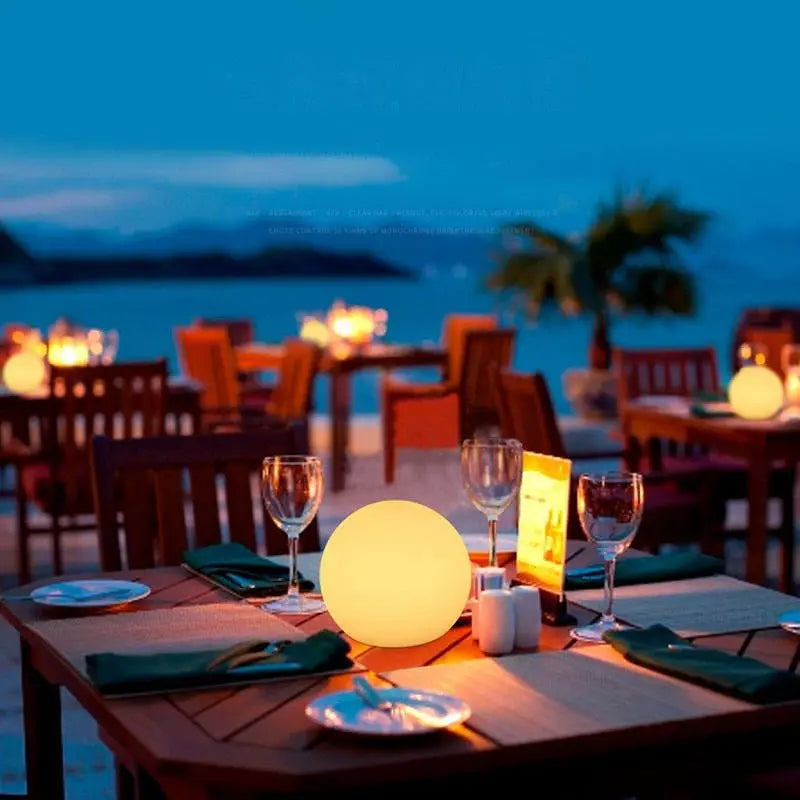 Leuchtende LED-Kugellampe auf einem gedeckten Holztisch eines Strandrestaurants am Abend mit Gläsern und Tellern.