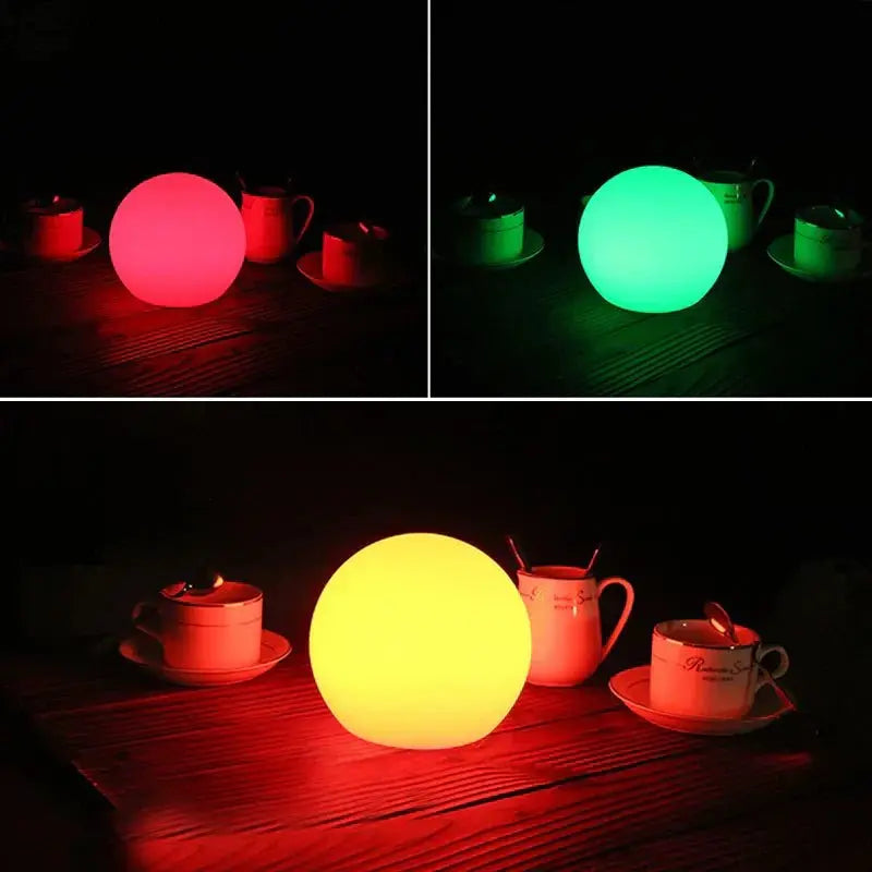 Drei Aufnahmen der LED-Kugellampe in Rot, Grün und Gelb, jeweils im Dunkeln neben Tassen auf einem Holztisch.