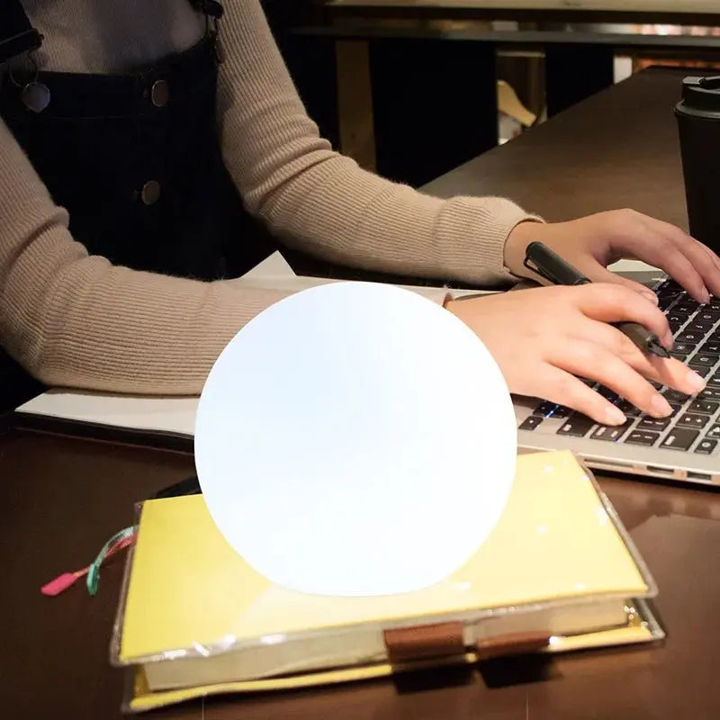 Hell weiß leuchtende LED-Kugellampe auf einem gelben Notizbuch neben einer Person, die an einem Laptop tippt.