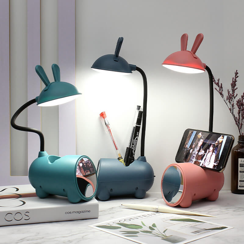 Drei leuchtende Hasenlampen in Grün, Dunkelblau und Rosa auf einem Schreibtisch mit Stiften und einem Smartphone im Halter.