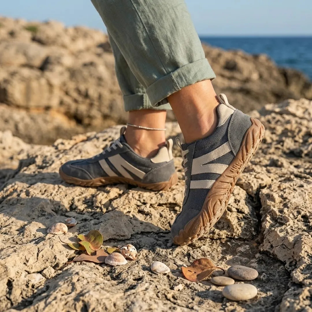Graue Barfußschuhe mit beigen Streifen, getragen auf felsiger Küste mit Muscheln und Kieselsteinen, Hosenbein hochgekrempelt. 2