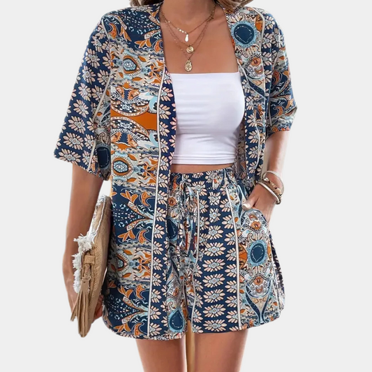 Frau in blau-orange gemustertem Sommeranzug, weißem Top, mit Strohtasche und Schmuck.
