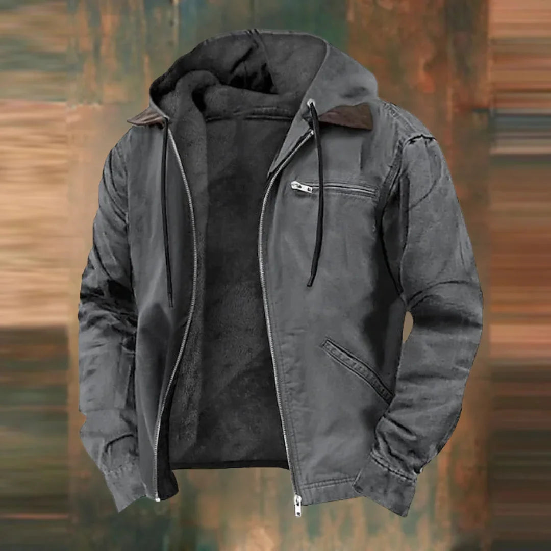 Die Jacke in Anthrazitgrau, offen mit Kapuze, Kordeln und dunklem Plüschfutter vor unscharfem Hintergrund.