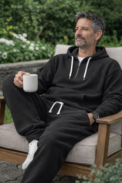 Mann im schwarzen Waffelstrick-Set sitzt mit einer Tasse in einem Holzsessel im Garten. 5