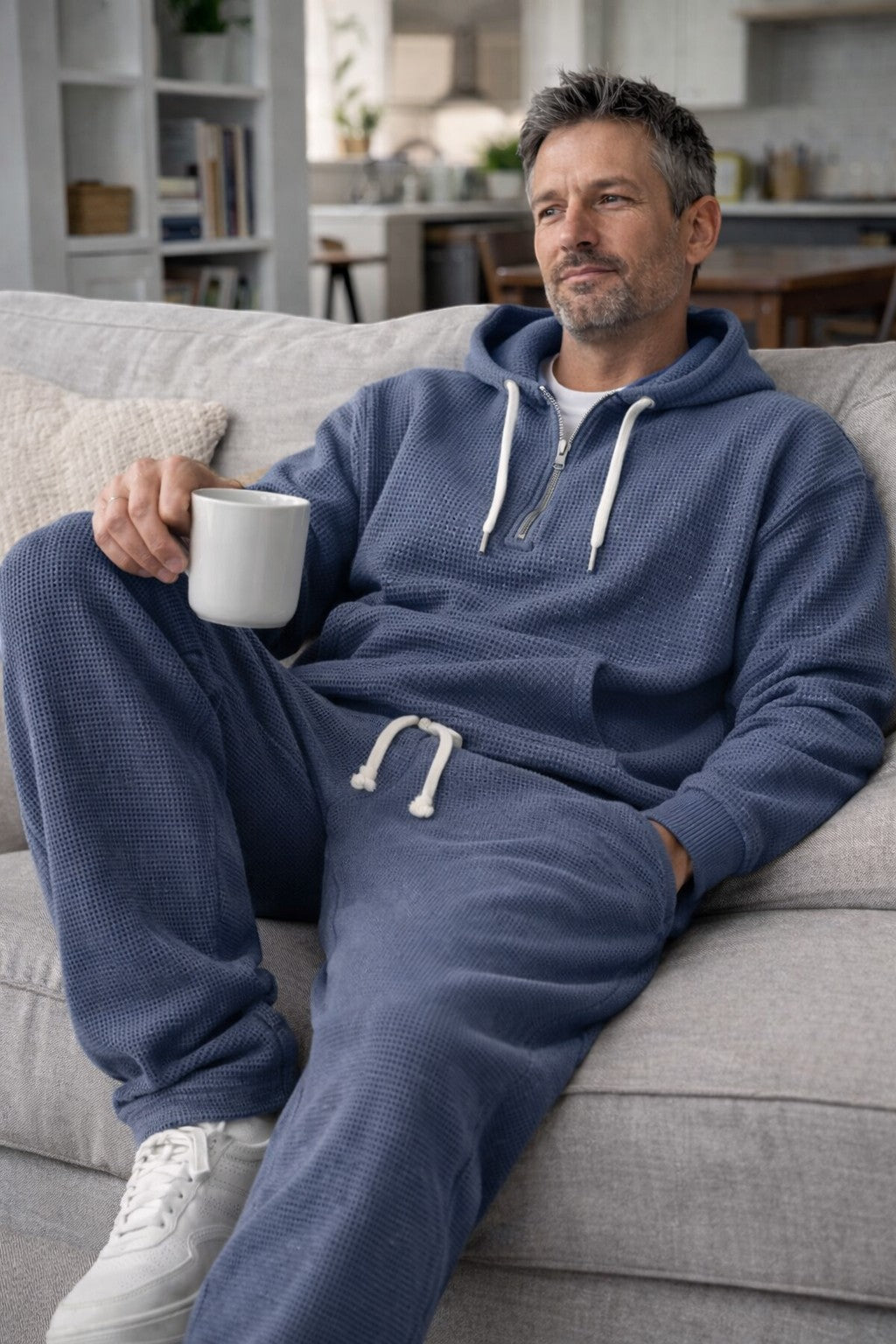 Mann im blauen Waffelstrick-Set entspannt mit einer Tasse auf einem hellgrauen Sofa. 4