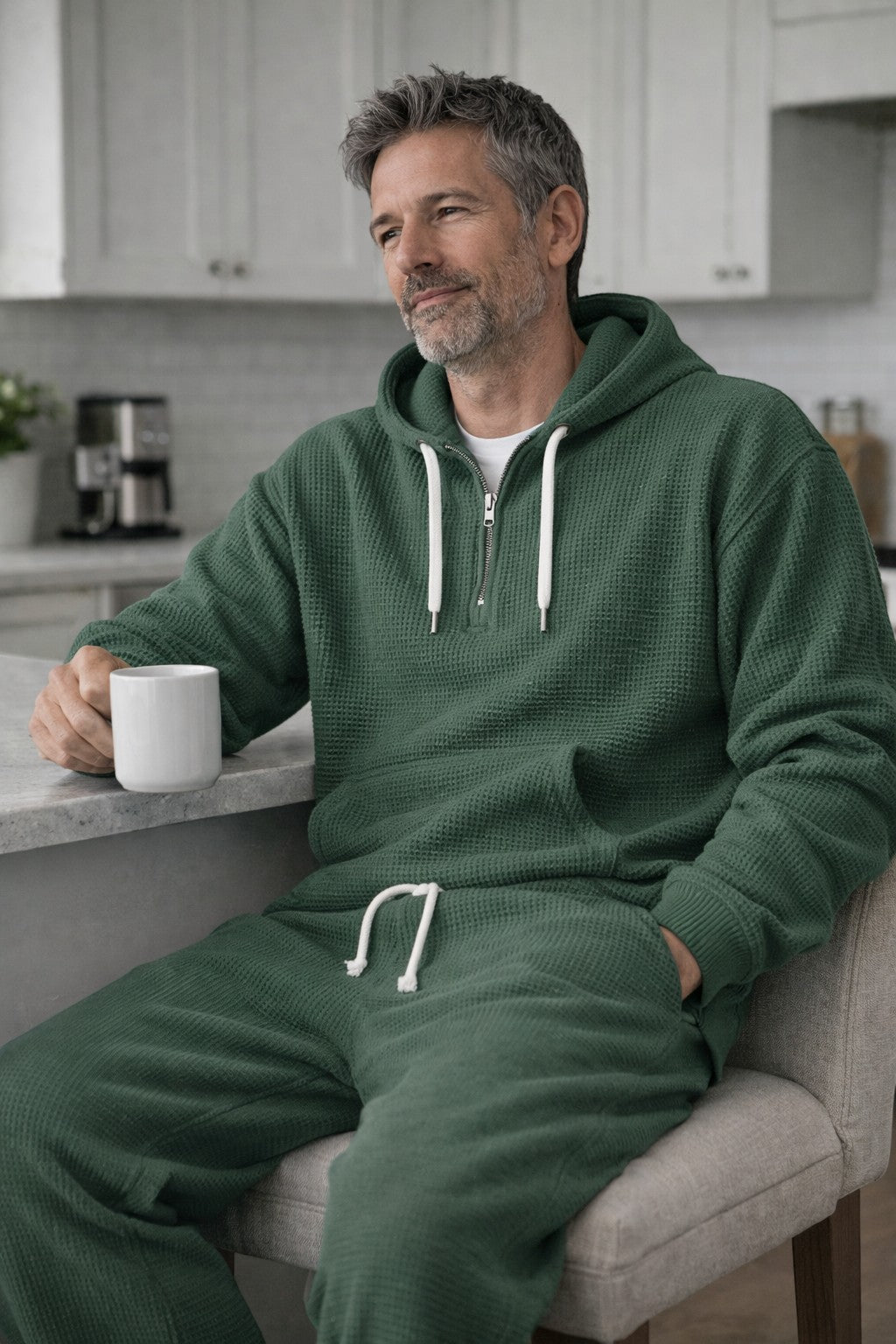 Mann in grünem Waffelstrick-Hoodie und passender Hose sitzt mit einer Tasse an einer Küchentheke. 3
