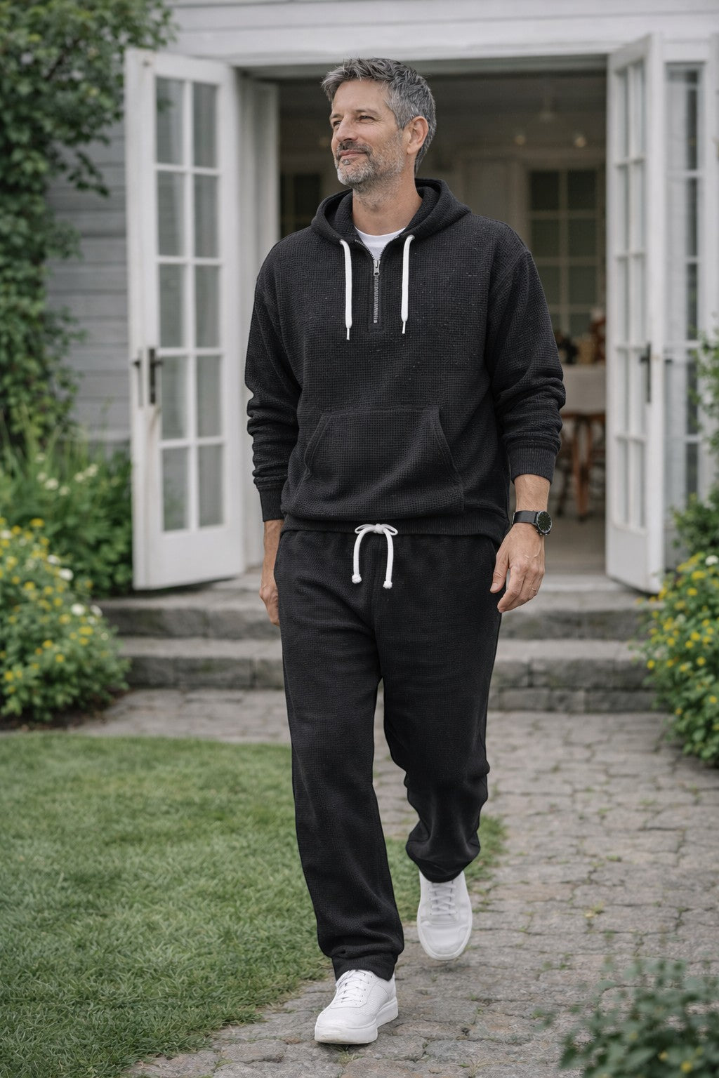 Mann in schwarzem Waffelstrick-Set mit Hoodie und Jogginghose geht in einem Garten vor einem weißen Haus spazieren. 1