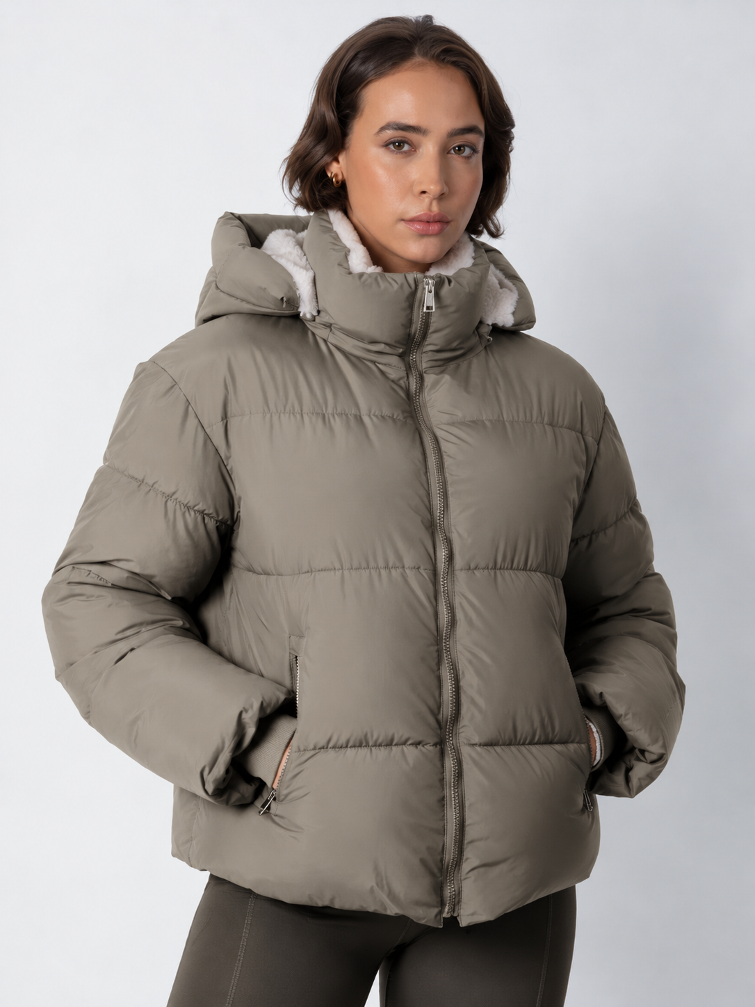 Model in geschlossener graubeiger Steppjacke mit Kapuze und Fleecefutter, Hände in den Taschen 18