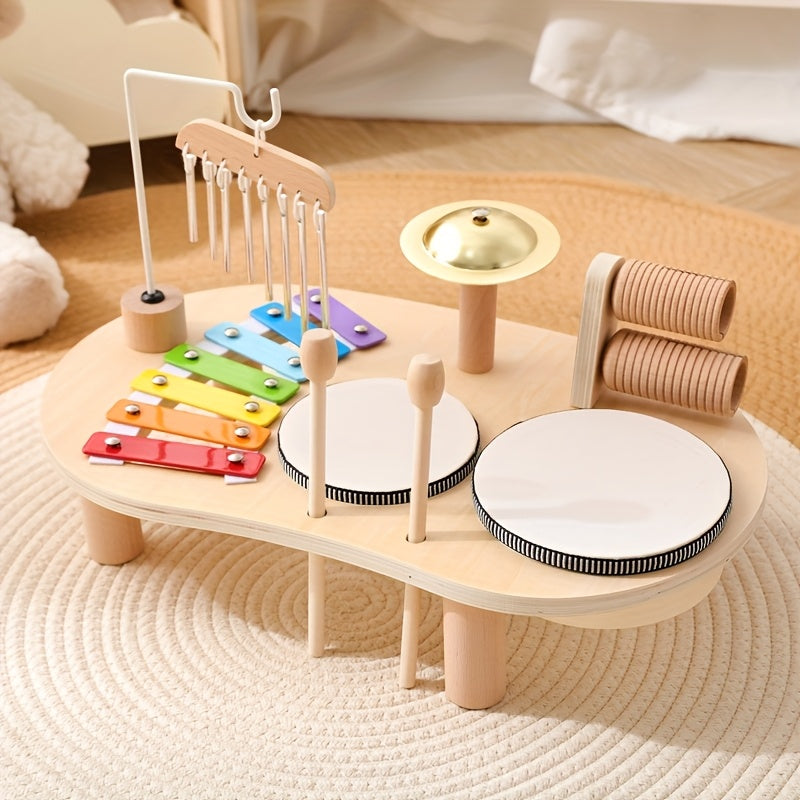 Holz-Musiktisch mit buntem Xylophon, Klangstäben, Becken, zwei Trommeln und Rassel-Walzen auf einem hellen Teppich, schräg von vorn. 1