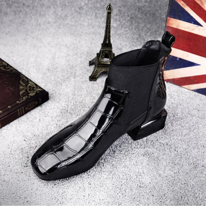 Schwarzer glänzender Stiefel mit Krokoprägung und elastischem Einsatz, schräg von vorn auf grauem Teppich neben Eiffelturm-Figur fotografiert. 1