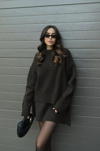 Model in dunkelbraunem Strick-Zweiteiler lehnt an einer Wand, eine Hand in der Rocktasche, schwarze Tasche in der anderen. 13