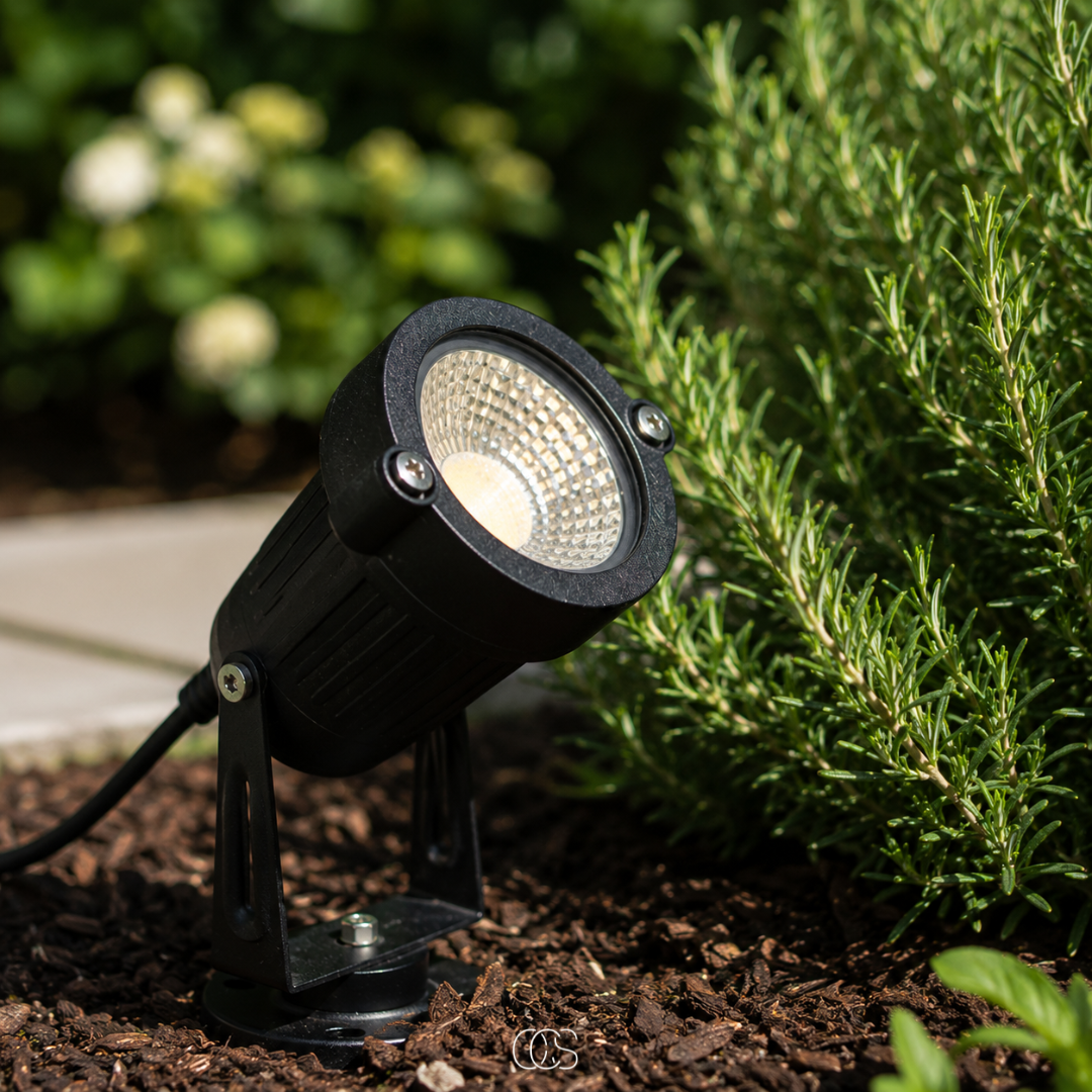 Schwarzer LED-Gartenstrahler mit Erdspieß aus Metall in detail auf Rindenmulch neben Rosmarin und Weg.