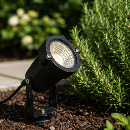 Schwarzer LED-Gartenstrahler mit Erdspieß aus Metall in detail auf Rindenmulch neben Rosmarin und Weg.