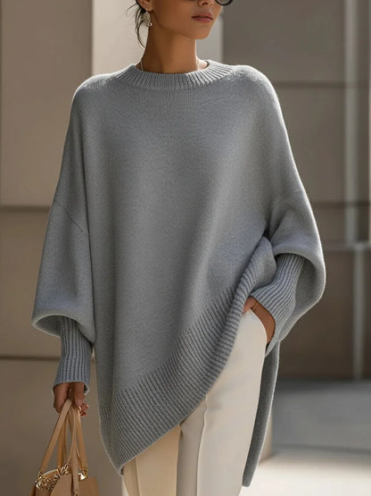 Grauer Oversize-Pullover mit gerippten Bündchen und asymmetrischem Saum, vom Model mit weißer Hose getragen. 9