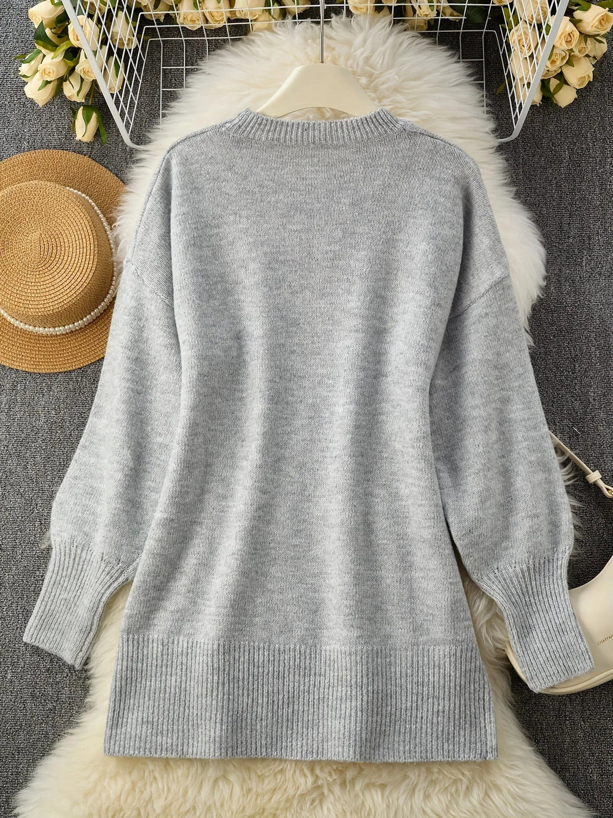 Grauer Strickpullover flach auf einem weißen Fellteppich ausgelegt, Rückseite mit Rippsaum, umgeben von Strohhut, Rosen und Tasche. 10