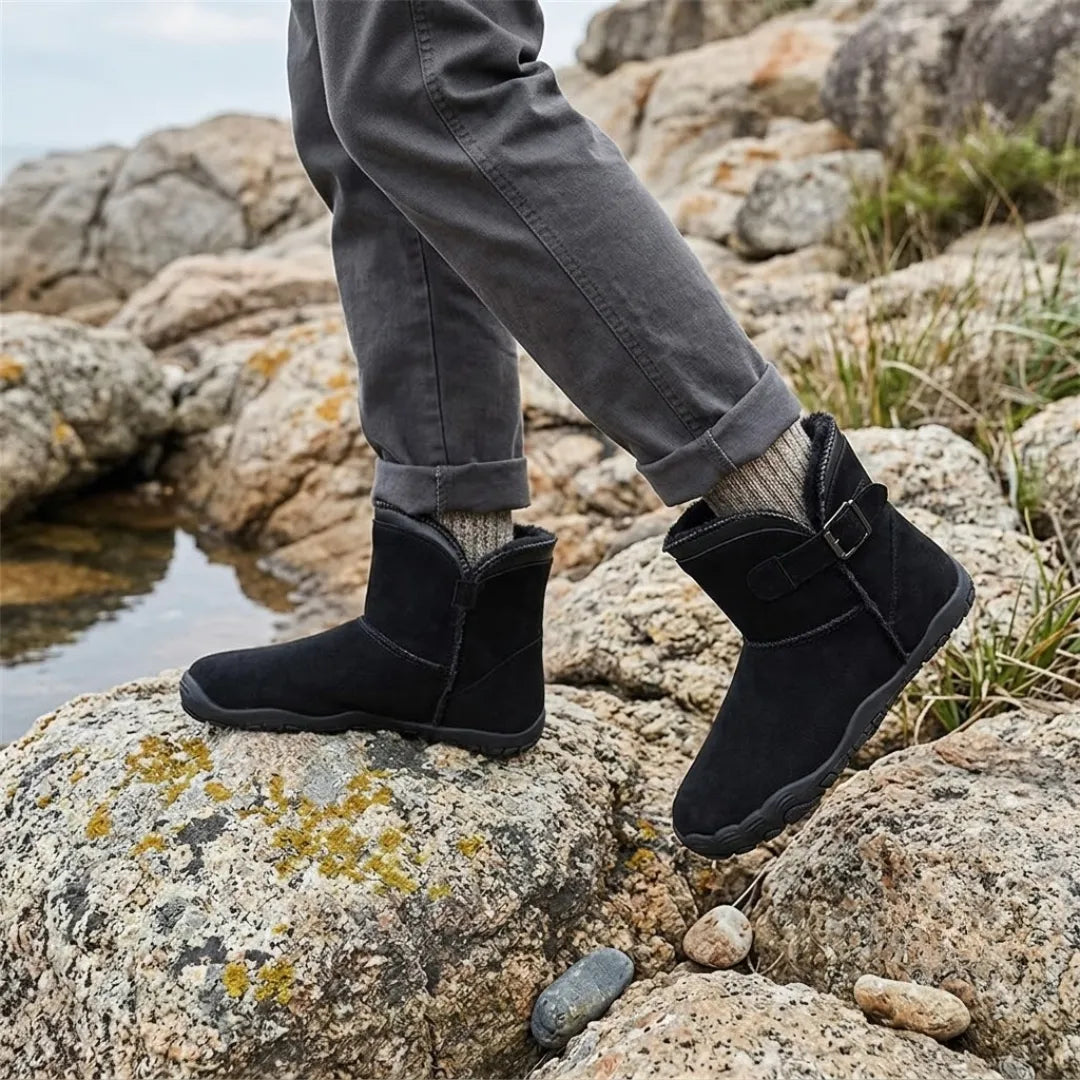 Schwarze Stiefel mit Schnalle, getragen mit Wollsocken und grauer Hose auf Felsen am Wasser 11