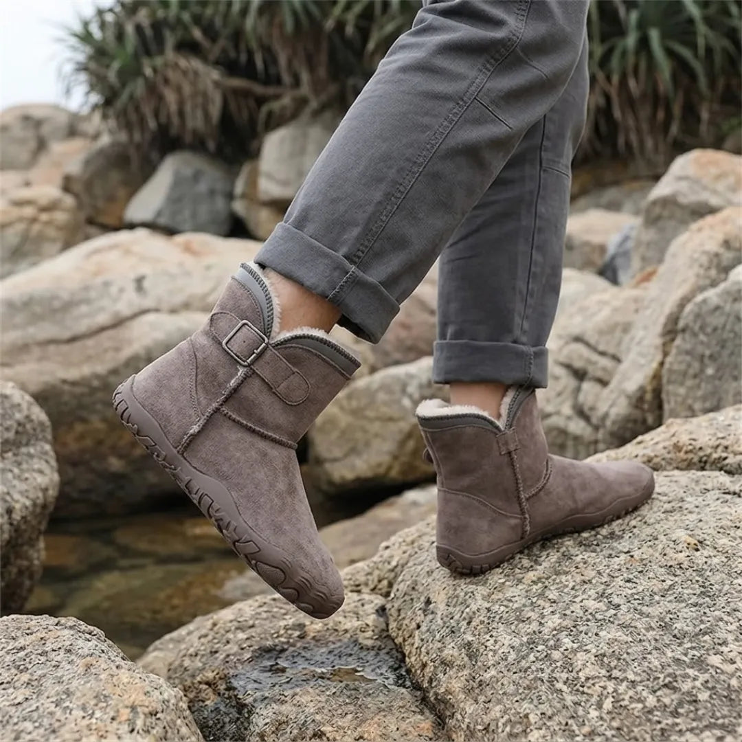 Graubraune Stiefel an den Füßen eines Trägers in grauer Hose, der auf Felsen am Wasser steht 38