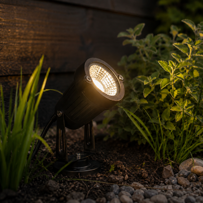 Leuchtender schwarzer LED-Gartenstrahler mit Erdspieß aus Metall im Beet vor dunkler Holzwand.