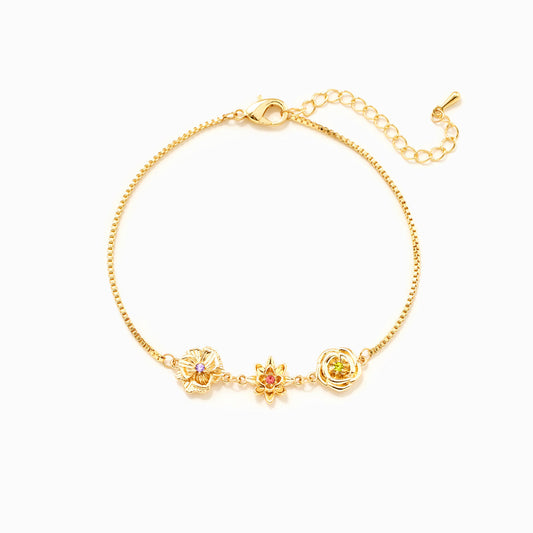 Goldfarbenes Gliederarmband mit drei Blumenanhängern und kleinen bunten Steinen, flach auf weißem Hintergrund. 1