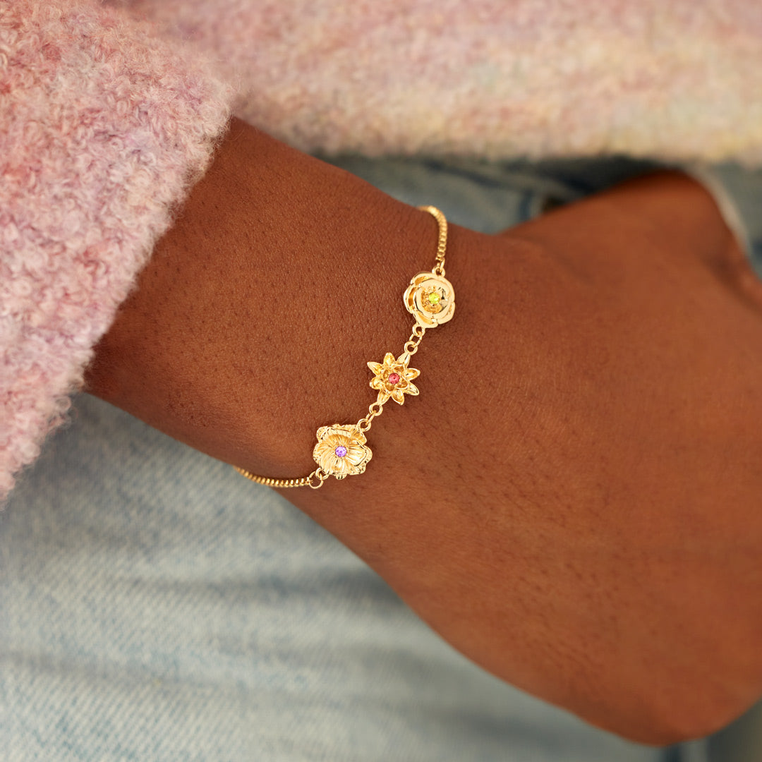 Am Handgelenk getragenes goldenes Blumenarmband mit drei Anhängern, dazu ein rosa Flauschpulli und hellblaue Jeans. 9