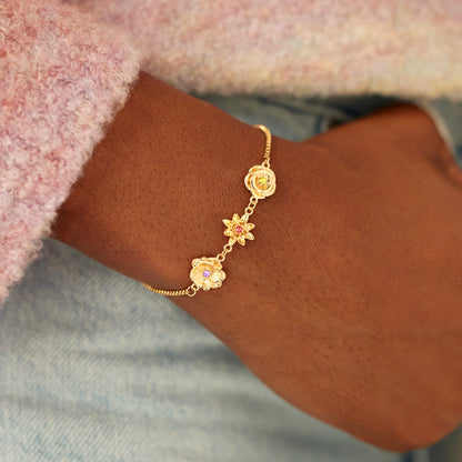 Am Handgelenk getragenes goldenes Blumenarmband mit drei Anhängern, dazu ein rosa Flauschpulli und hellblaue Jeans. 9