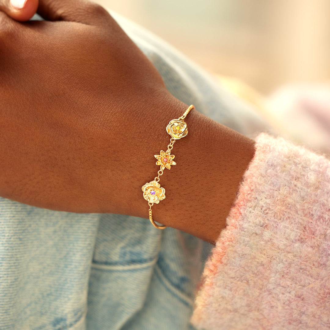Handgelenk mit dem goldenen Blumenarmband von der Seite, im Hintergrund ein rosa Flauschärmel und eine Jeansjacke. 10