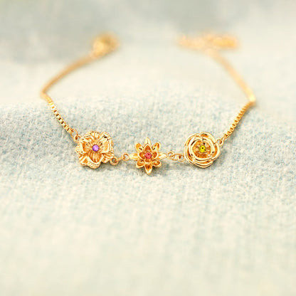 Goldenes Blumenarmband mit drei bunten Steinen auf hellblauem, flauschigem Stoff. 15
