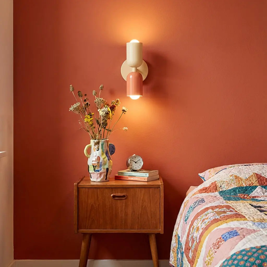Leuchtende Wandlampe in Creme und Lachsrosa an terrakottafarbener Wand über einem Holz-Nachttisch mit bunter Vase und Trockenblumen. 1